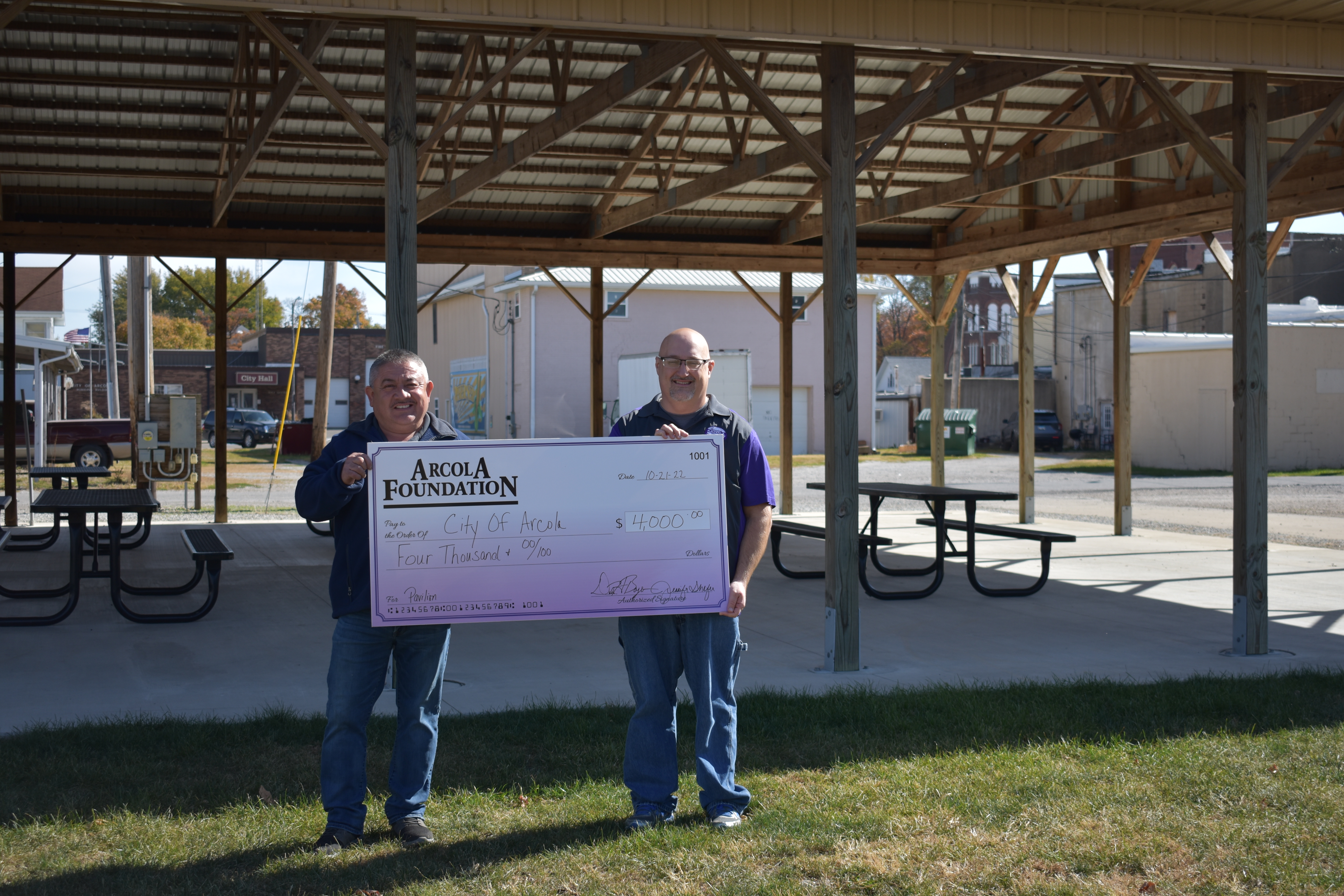 Two men holding a large check while standing in front of a pavilion outdoors